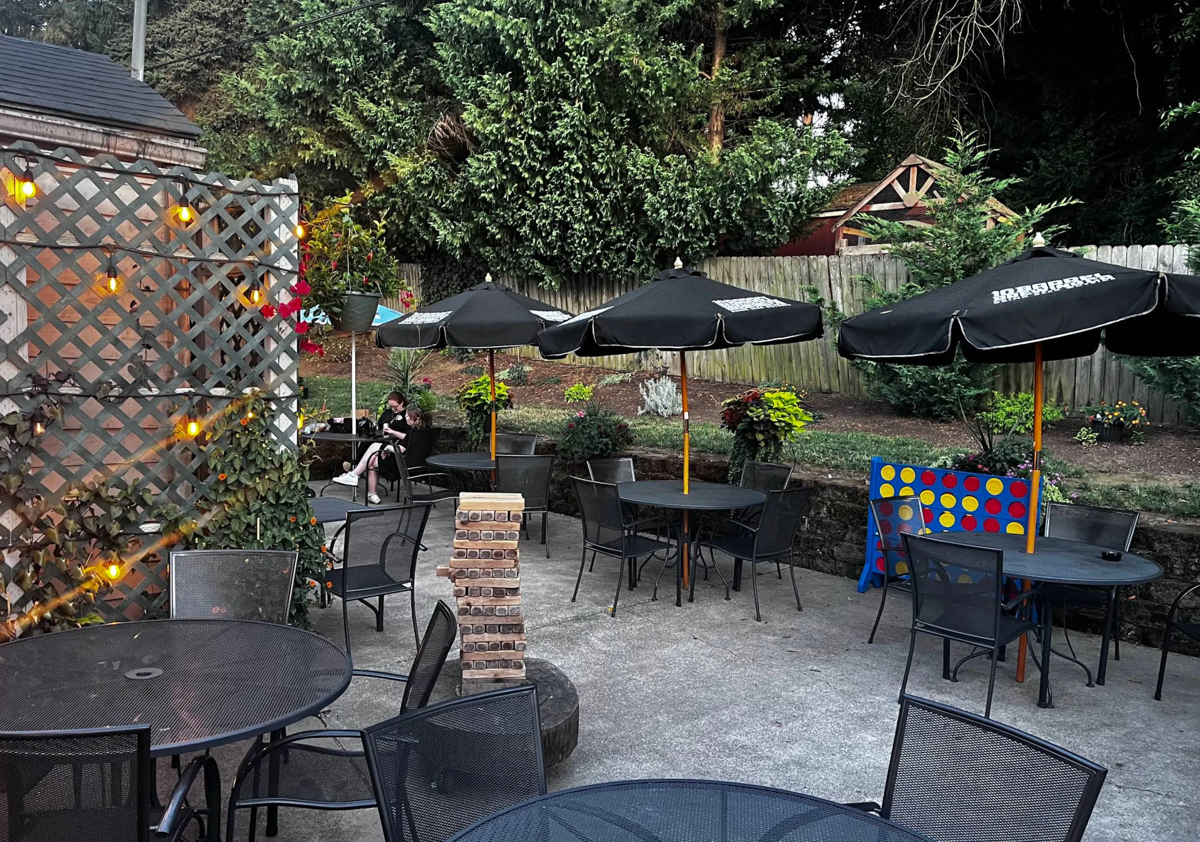 The outdoor patio at dusk — umbrella tables and string lights with giant Jenga and Connect Four set up under the trees.